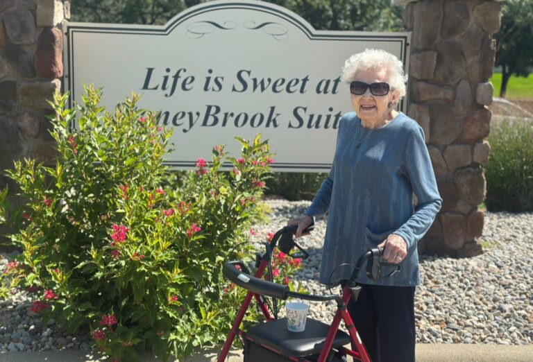 Elderly woman with a walker standing in front of a sign that says "Life is Sweet in StoneyBrook Suites"